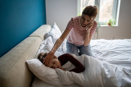 Photo for Worried young woman mother calling to pediatrician while sitting near sick son on bed. Stressed mom holding smartphone talking to doctor, feeling worried about child health, checking kids temperature - Royalty Free Image