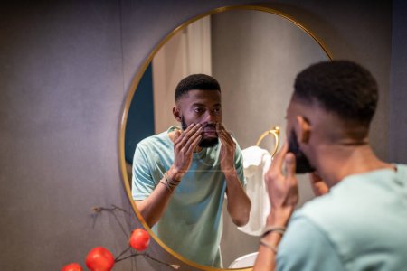Foto de Hombre africano joven preocupado infeliz mirando en el espejo tocando arrugas en la cara, tipo negro de 30 años de pie en el baño examinando la piel, preocupándose por el envejecimiento. Concepto de hombres y cuidado de la piel - Imagen libre de derechos