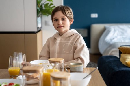 Foto de Retrato de un niño europeo feliz desayunando en casa. Sonriente niño colegial sentado en la mesa de la cocina por la mañana. Niño interesado mirando la cámara comiendo copos de maíz antes de la escuela - Imagen libre de derechos