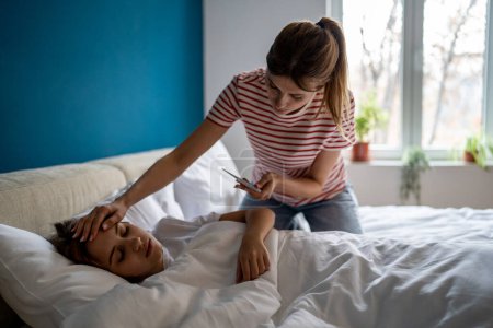 Photo for Worried caucasian mother measuring temperature of sick teen daughter lying in bed at home, caring mom parent holding thermometer touching head of unhealthy preteen child. Children and colds - Royalty Free Image
