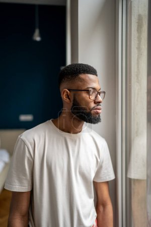 Photo for Upset young African man wearing white t-shirt standing at home looking out window and pondering, having problems in personal life. Frustrated sad black guy feeling down. Men and depression - Royalty Free Image