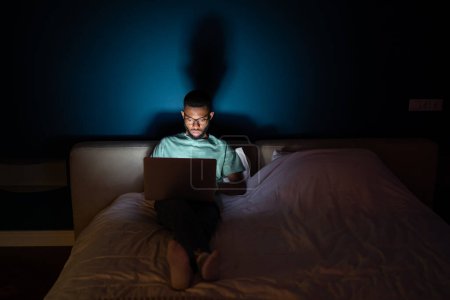 Photo for Black man works on laptop lying in bed at night in dark looks attentively at screen. Serious african american guy doing his job in bedroom at home. Workaholic, overwork and overjob concept. - Royalty Free Image