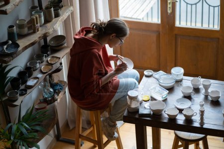 Foto de Inmersiva en el proceso de la mujer artesana sentada en el estudio de cerámica trabajando en la creación de vajilla única. Patrón de pinturas femeninas concentradas en placa de arcilla natural con pincel para la venta en la tienda de cerámica - Imagen libre de derechos