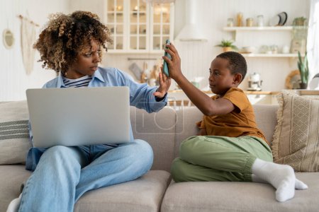 Photo for African american mom strictly looks at son with smartphone works on laptop sits on couch at home. Boy prevents mother to do job. Working mum, motherhood and career, distant remote work concept. - Royalty Free Image