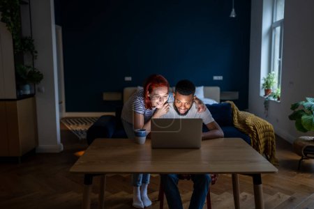 Photo for Happy young married multiracial couple using laptop together, planning summer vacation or shopping online. Caucasian girl and African American guy choosing movie to watch in evening at home - Royalty Free Image