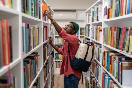 Photo for Nerd student african american man choosing book in university library taking it from bookshelf. Wistful guy hipster with backpack studying college. Education, giving new profession, learning concept. - Royalty Free Image