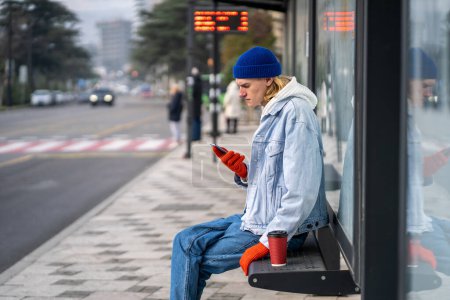 Photo for Stylish guy waiting bus sitting on bus stop bench in downtown with coffee cup browsing smartphone. Public transport problem concept. Man searching bus routes information, transport schedule timetable. - Royalty Free Image