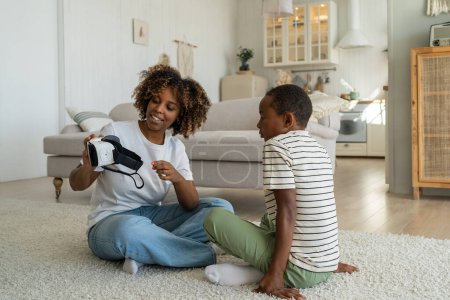 Photo for Satisfied pleased African American mother hold virtual reality glasses for smartphone together with child boy. Content black woman show schoolboy son VR headset for 360 video immersion game at home - Royalty Free Image