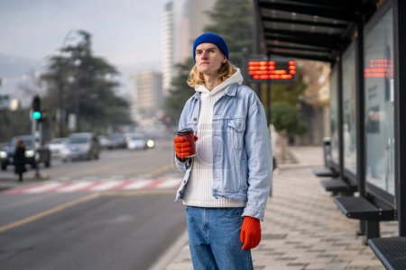 Müder Typ Hipster, der an einer Bushaltestelle in der Innenstadt steht, hält eine Kaffeetasse in der Hand. Problem ÖPNV im Stadtkonzept. Moderne, menschenfreundliche Stadtumgebung. Getränke auf der Straße genießen.