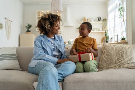 Photo for Caring loving African American mom greeting surprised small boy with present gift. Kind black woman give on birthday celebrate wrapped box with red ribbon to anticipating son sitting on couch at home - Royalty Free Image