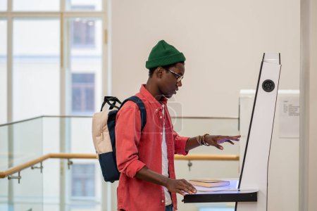Foto de Student African man using self-service electronic terminal to pay for goods in store without salesperson. Self-sufficient black guy standing indoors at college using touch screen vending machine - Imagen libre de derechos