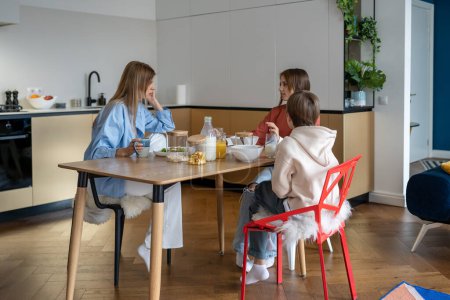 Photo for Single mother with children sit at kitchen table have breakfast before school. Family morning routine. Teen daughter talk mom and younger brother about plan of college study. Healthy eating together - Royalty Free Image