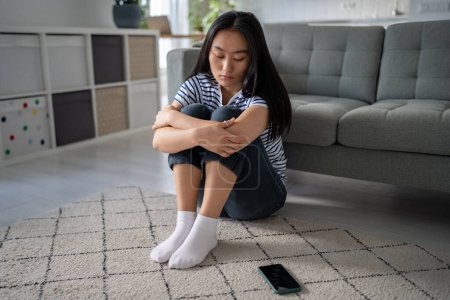 Photo for Unhappy depressed Asian girl sitting on floor at home looking at smartphone waiting for call from boyfriend, feeling sad and heartbroken after receiving break up text message, cant forget ex - Royalty Free Image