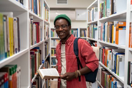 Photo for Smiling african american man student holding book in university library standing between bookshelves. Life in campus, education, self-development reading books studying learning researching concept. - Royalty Free Image