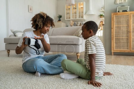 Photo for VR for kids. Modern African American family mother and son sitting on floor with virtual reality headset, parent mom holding AR smart glasses introducing child to augmented reality technology - Royalty Free Image