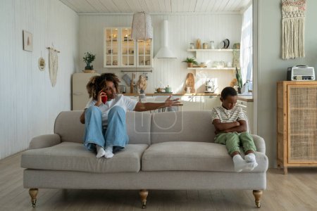 Photo for Annoyed angry African American mother sitting with offended upset son on sofa at home, talking on phone, discussing child behavior with spouse. Mom and kid conflict. Parents, children and discipline - Royalty Free Image