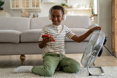Téléchargez les photos : Joyeux petit garçon souriant afro-américain regarde la caméra se trouve avec téléphone portable dans les écouteurs sur le refroidissement du tapis tenant ventilateur de sol électrique. Écolier satisfait écouter de la musique sur smartphone à la maison - en image libre de droit