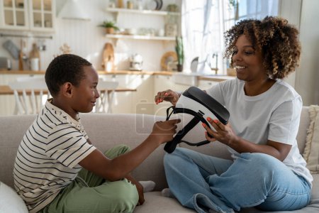 Téléchargez les photos : Mère afro-américaine aidant son fils avec des lunettes de réalité virtuelle à la maison, aidant à utiliser le casque en toute sécurité, mère heureuse et enfant assis ensemble sur le canapé explorer le monde virtuel. Expérience de l'enfant et vr - en image libre de droit