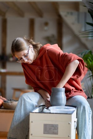 Téléchargez les photos : Créatrice artisanale enseignante de cours de céramique façonnant cruche se trouve près de la roue des potiers en atelier. Fille professionnelle travaille sur studio en céramique aider à apprendre à faire de la vaisselle à la main pour la vente en ligne - en image libre de droit