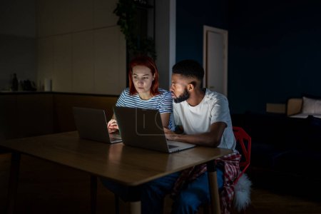 Téléchargez les photos : Jeune couple interracial homme et femme regardant ordinateur portable discuter entreprise familiale en ligne, travailler ensemble la nuit, assis à table dans la pièce sombre à la maison. Concept d'entrepreneuriat familial - en image libre de droit