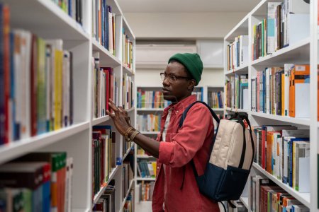 Photo for Interested Black student man choosing book for reading in bookstore. Focused young African American guy searching materials for educational research in college library. Literature and education - Royalty Free Image