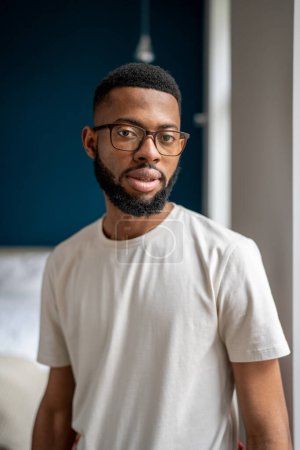 Téléchargez les photos : Plan vertical d'un jeune homme noir millénariste portant un t-shirt basique et des lunettes regardant la caméra, portrait domestique rapproché d'un jeune homme afro-américain posant dans un appartement. - en image libre de droit