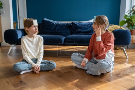 Photo for Two kids boy and girl friends playing Who Am I guessing game while sitting floor in living room. Children brother and sister play together, siblings at home. Indoor entertaining activities for kids - Royalty Free Image