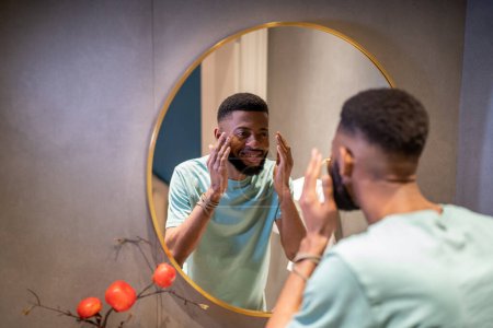 Téléchargez les photos : Afro-Américain regardant reflet dans le miroir et souriant, heureux mec noir debout dans la salle de bain laver la peau du visage avec de l'eau le matin, se sentant satisfait de son apparence, accepter le vieillissement - en image libre de droit