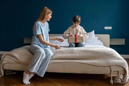 Photo for Happy little kid opening birthday present from mom, excited smiling boy sitting on bed with mother unwrapping wrapped gift box. Loving parent congratulating child at home - Royalty Free Image