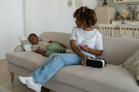 Téléchargez les photos : Petit enfant afro-américain dormant sur le canapé après une longue utilisation du casque VR à la maison, aimant mère attentionnée parent tenant casque de réalité virtuelle regardant fatigué fils endormi couché à proximité. - en image libre de droit