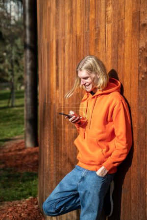 Foto de Sonriente rubia delgado hipster chico lee mensaje divertido en el teléfono de chat con amigos se para cerca de la valla de madera en el parque natural. Las redes sociales en línea reemplazan la comunicación real con virtual a pie. - Imagen libre de derechos