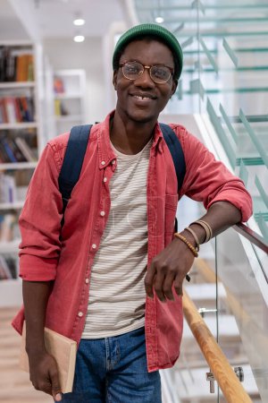 Foto de Sonriendo satisfecho afroamericano estudiante en gafas de pie en la biblioteca de la universidad mirar a la cámara. Retrato feliz complacido chico negro hipster con libros de texto de pie en la universidad después de la conferencia - Imagen libre de derechos