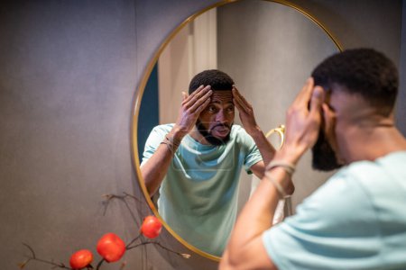 Foto de Infeliz hombre afroamericano de 30 años tocando la piel de la cara mirando el espejo durante la rutina diaria de la mañana en el baño, chico negro se siente preocupado por las primeras arrugas, pensando en el tratamiento cosmético anti-edad - Imagen libre de derechos