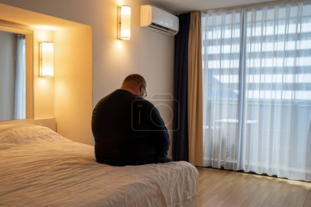 Photo for Obesity man sadly sitting on bed on vacation in hotel alone without friends having midlife crisis, rear view. Overweight insecure male in depression suffers from loneliness, having life problems. - Royalty Free Image