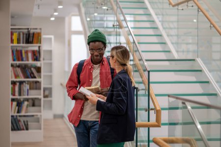 Photo for Smiling diverse multiethnic student couple talk in university campus library share information about lecture. Cheerful african american guy speak with caucasian girl holding textbook near glass ladder - Royalty Free Image