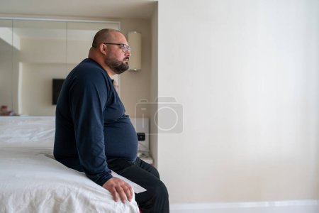Photo for Calm sleepy man waking up in morning at home sitting in bed ready to start new day, side view. Restful overweight bearded young male in bedroom. Early rise, healthy sleeping and lifestyle concept. - Royalty Free Image