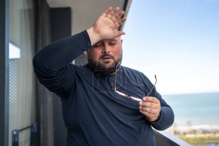 Photo for Overweight problem. Exhausted heavy man touch forehead feeling blood pressure from heat weather. Unhealthy tired fat male with increased perspiration standing on balcony to difficult breathe air - Royalty Free Image