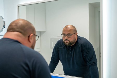 Photo for Confused upset overweight middle aged man looking at mirror in bathroom at home. Bearded male in eyeglasses dissatisfied with appearance. Midlife crisis, psychological problems, depression concept. - Royalty Free Image