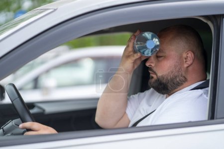 Exhausted overweight man driver suffering from heat inside car with broken air conditioner touch bottle of water to forehead closed eyes try cool. Male feel sick headache from hot air temperature 