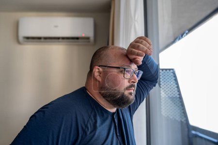 Photo for Overweight man hiding at home with working air conditioner in hot weather looking at window. Bearded exhausted from heat male in apartment suffering from high air temperature having healthy problems. - Royalty Free Image
