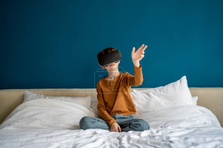 Téléchargez les photos : Enfant inspiré faisant l'expérience de lunettes de réalité virtuelle immersives dans un monde futuriste. Intéressé petit écolier à la recherche de lunettes VR pour smartphone jouer dans le jeu vidéo toucher l'air de la main assis sur le lit - en image libre de droit