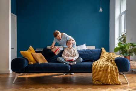 Photo for Young happy loving mother closing eyes of child congratulating little boy son with happy birthday at home, smiling mom giving wrapped gift box to kid sitting on sofa with smartphone. - Royalty Free Image