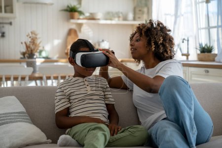 Téléchargez les photos : Afro-américaine maman moderne aidant petit garçon fils à mettre sur casque de réalité virtuelle tout en profitant des loisirs ensemble le week-end, mère présentant enfant avec une technologie impressionnante de jeu VR - en image libre de droit