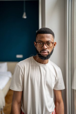 Téléchargez les photos : Sérieux homme noir millénial portant un t-shirt de base et des lunettes regardant la caméra, gros plan portrait domestique de jeune homme afro-américain propriétaire posant dans l'appartement. - en image libre de droit
