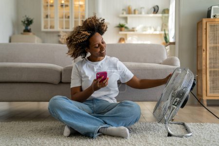 Photo for Summer heat at home. Happy curly African American woman sitting with smartphone on floor enjoying cool fresh air blowing from electric fan, chatting on mobile phone relaxing in front of air cooler - Royalty Free Image