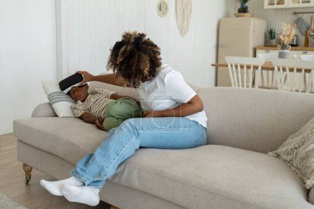 Téléchargez les photos : Maman enlève le casque de réalité virtuelle d'un adolescent accro assis sur un canapé à la maison. Garçon dormir dans des lunettes vr jouer à des jeux regarder un film. Sur l'utilisation de gadgets, jeu vr addiction, concept épuisé. - en image libre de droit