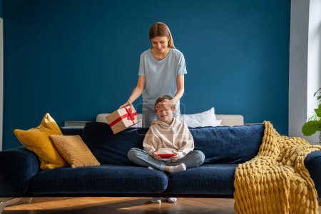 Photo for Loving mother parent making birthday surprise to kid son holding wrapped gift box, mom covering eyes with hand of excited smiling child boy sitting on sofa and waiting for present - Royalty Free Image