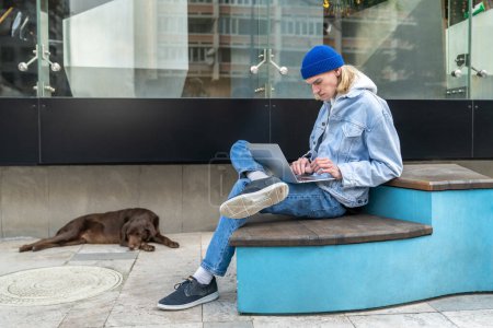 Téléchargez les photos : Homme hipster concentré pigiste travaillant sur ordinateur portable à l'extérieur, faire du travail en ligne tout en étant assis sur un banc avec chien à proximité. Millennial guy freelance avec animal de compagnie en plein air, en utilisant le wifi public pour le travail à distance - en image libre de droit