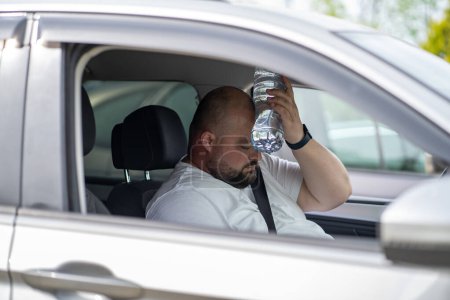 Photo for Exhausted overweight man driver suffer from blood pressure sits inside car hot weather puts bottle of water to forehead. Overheat male stopped after drive car in traffic jam feel trying to cool down - Royalty Free Image