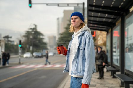 Téléchargez les photos : Millennial guy with blonde hair wearing casual denim clothes standing alone at public transport stop, young man with take away coffee cup waiting for bus outdoors. Transports et vie urbaine - en image libre de droit